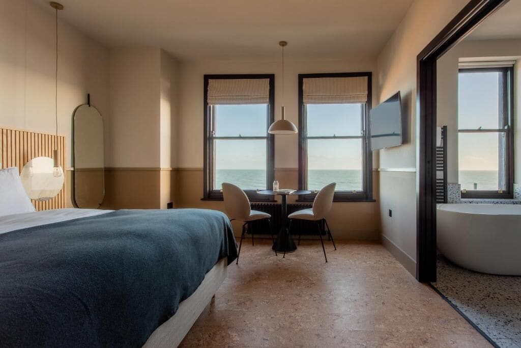 A hotel room with a bed, a dining table with chairs, a TV, and a view of the sea.