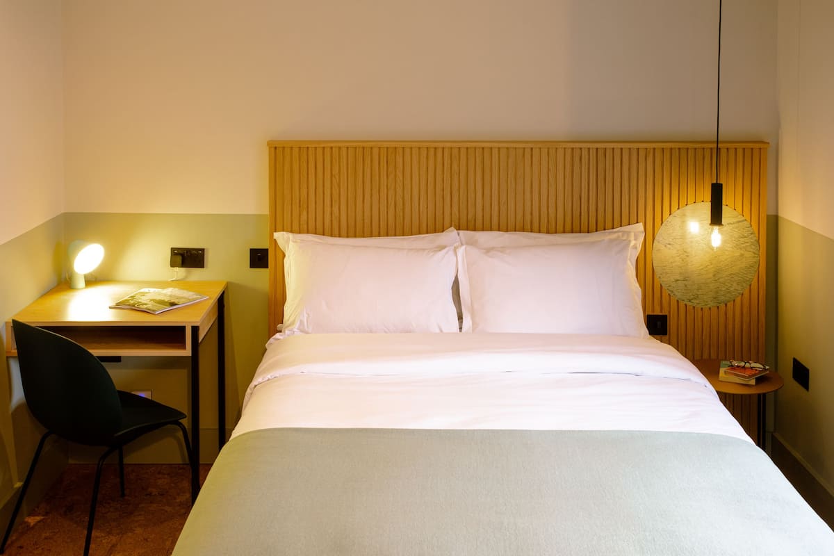 Hotellihuone, jossa on sänky, työpöytä tuolilla, lamppu ja pieni pöytä, jonka päällä on kirja.