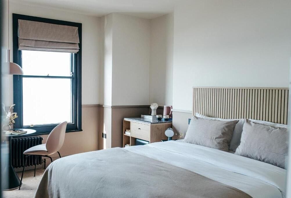 A hotel room with a large bed, a desk with a chair, a radiator, and a window with curtains.