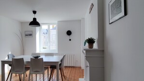 Et spiseområde med et bord, stole, en hængende sort lampe, et vindue med udsigt til bygninger, et ur og en plante på en hylde.