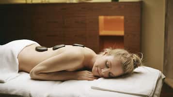 A person lying on a massage table with stones placed on their back, in a room with wooden furniture and a vase with flowers on a shelf.