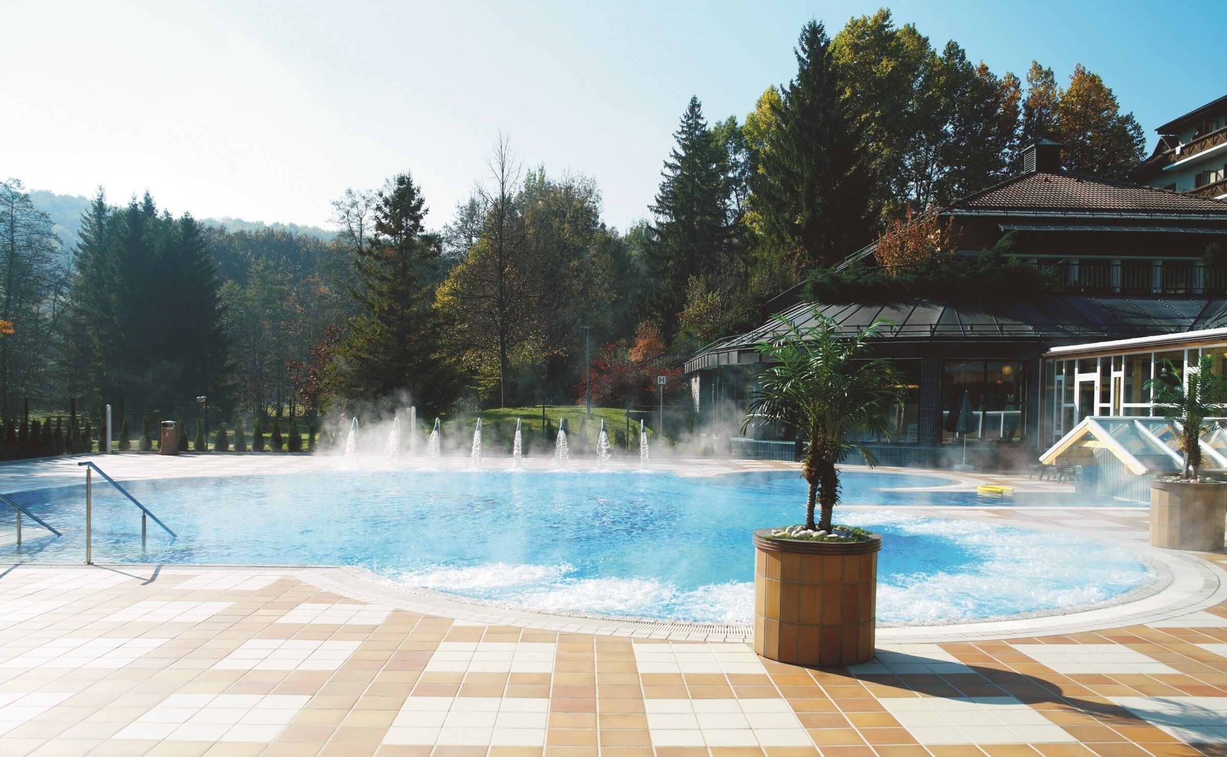 A swimming pool with a water fountain, surrounded by trees and a building with a sloped roof.