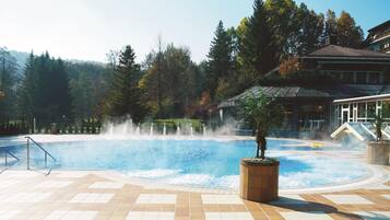 A swimming pool with a water fountain, surrounded by trees and a building with a sloped roof.