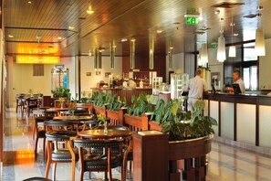 A hotel lobby with wooden flooring, multiple tables and chairs, potted plants, a reception desk, and a sign indicating the direction to the hotel rooms. - Hotel Toplice - Terme Krka (Smarjeske Toplice)
