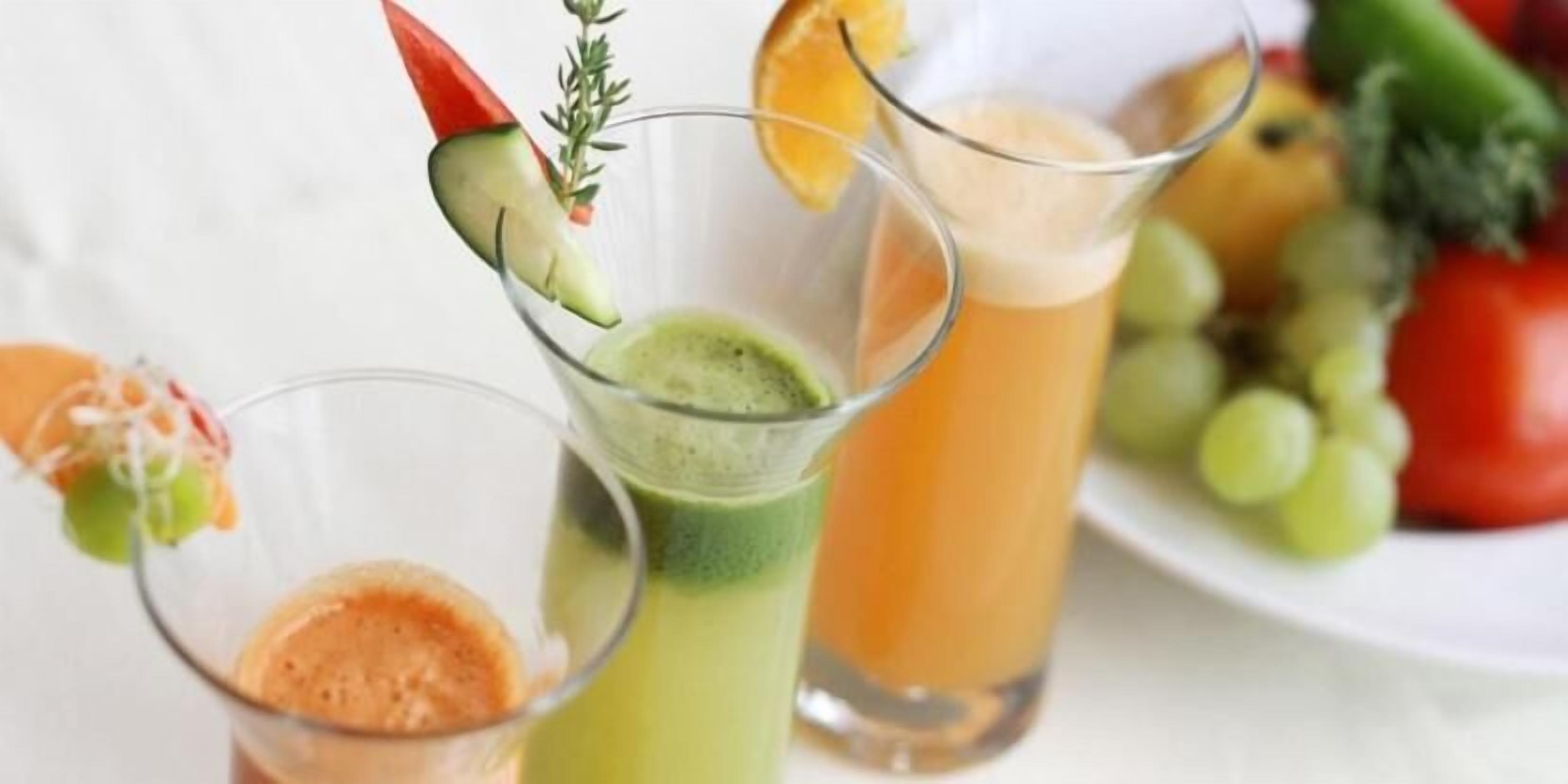 Tres vasos de bebidas de diferentes colores, un plato de frutas y un chile rojo como decoración.