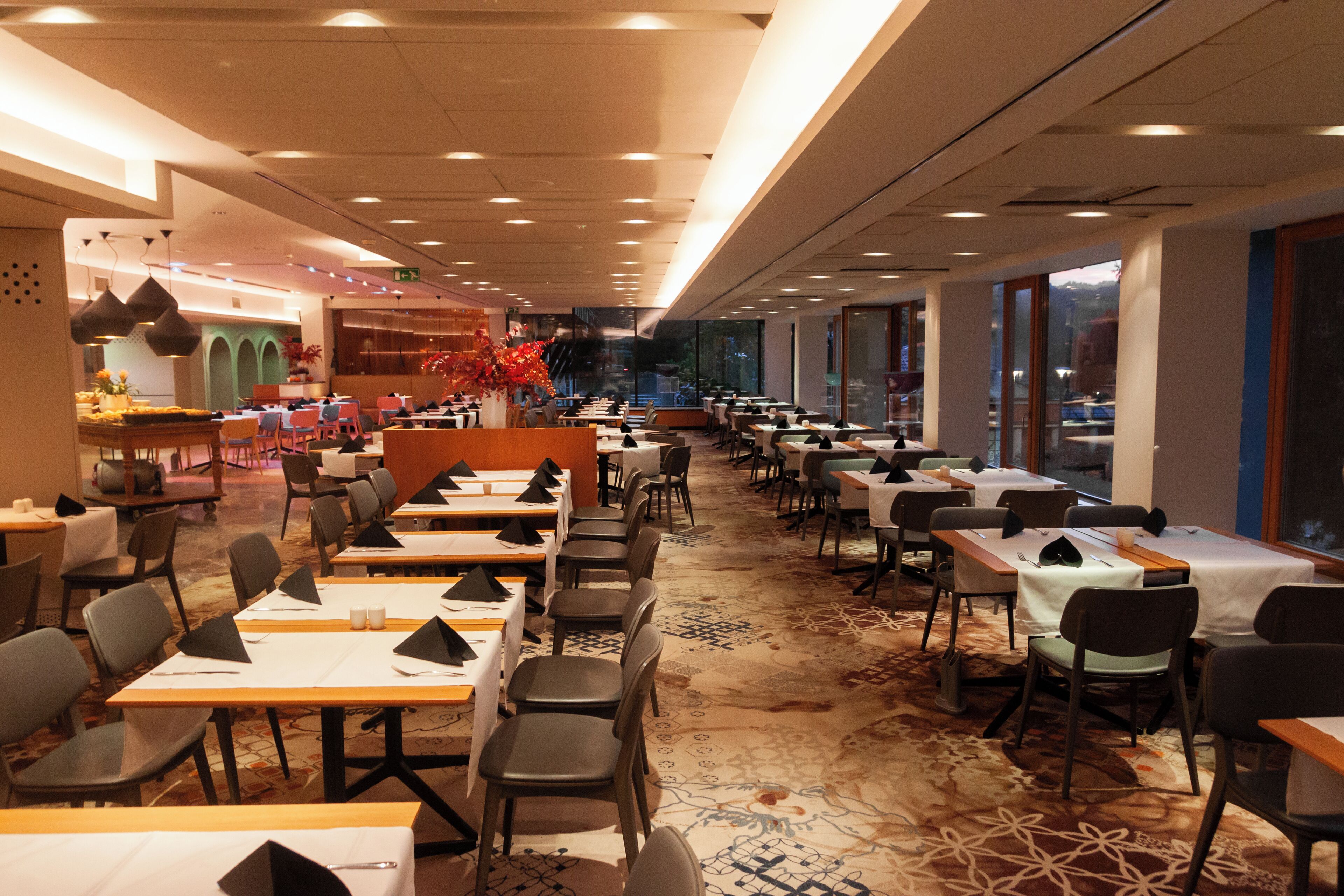 A spacious restaurant with multiple tables and chairs, a patterned carpet, and large windows offering a view.