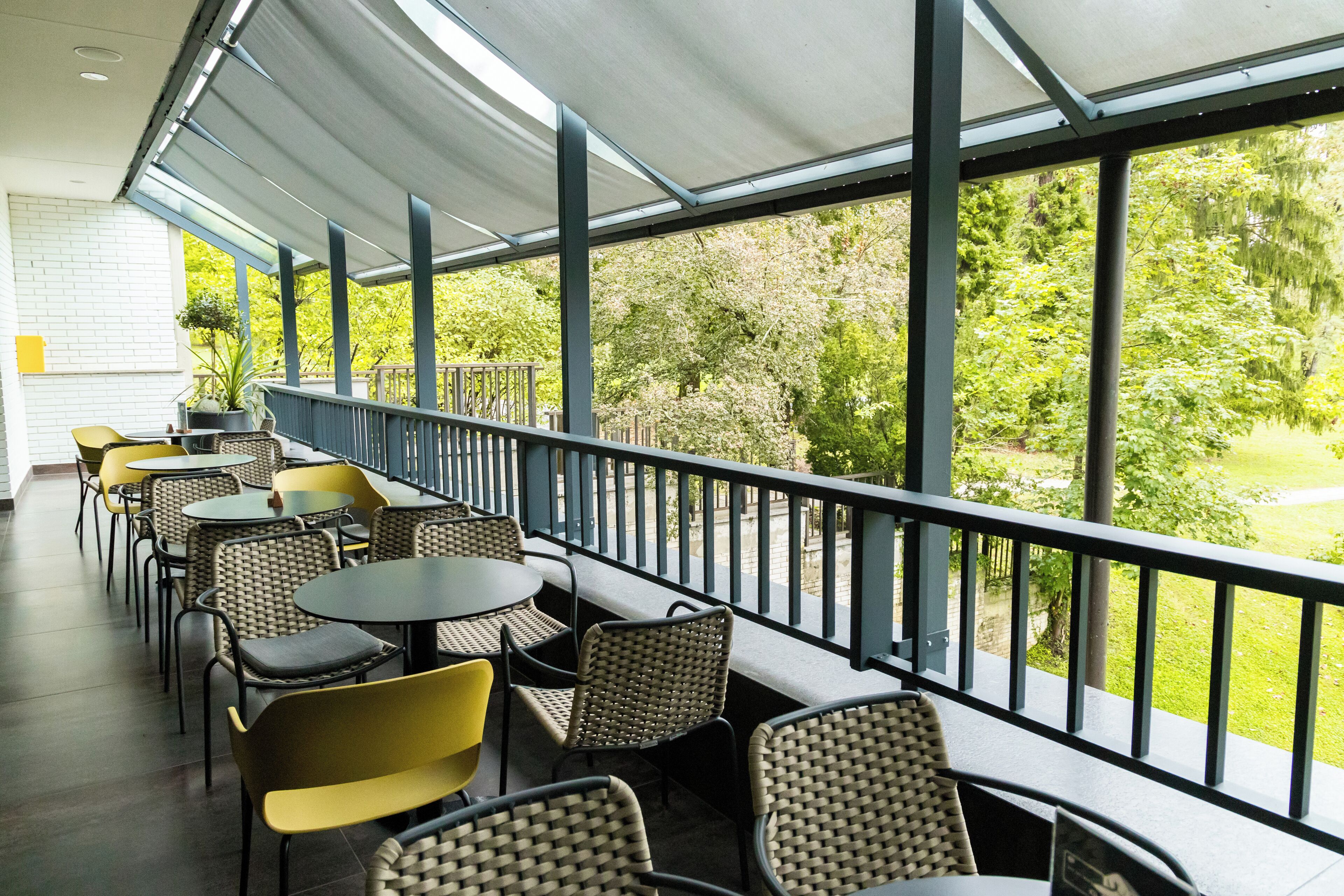 A covered outdoor seating area with tables and chairs, a railing, and a view of greenery.