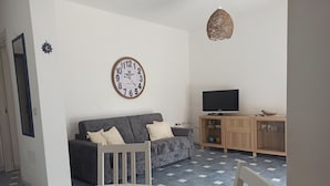 A living room with a grey sofa, a white table with a fruit basket, a wall clock, a TV on a wooden cabinet, and a hanging light fixture. - MaGi Holiday Home (Stintino)