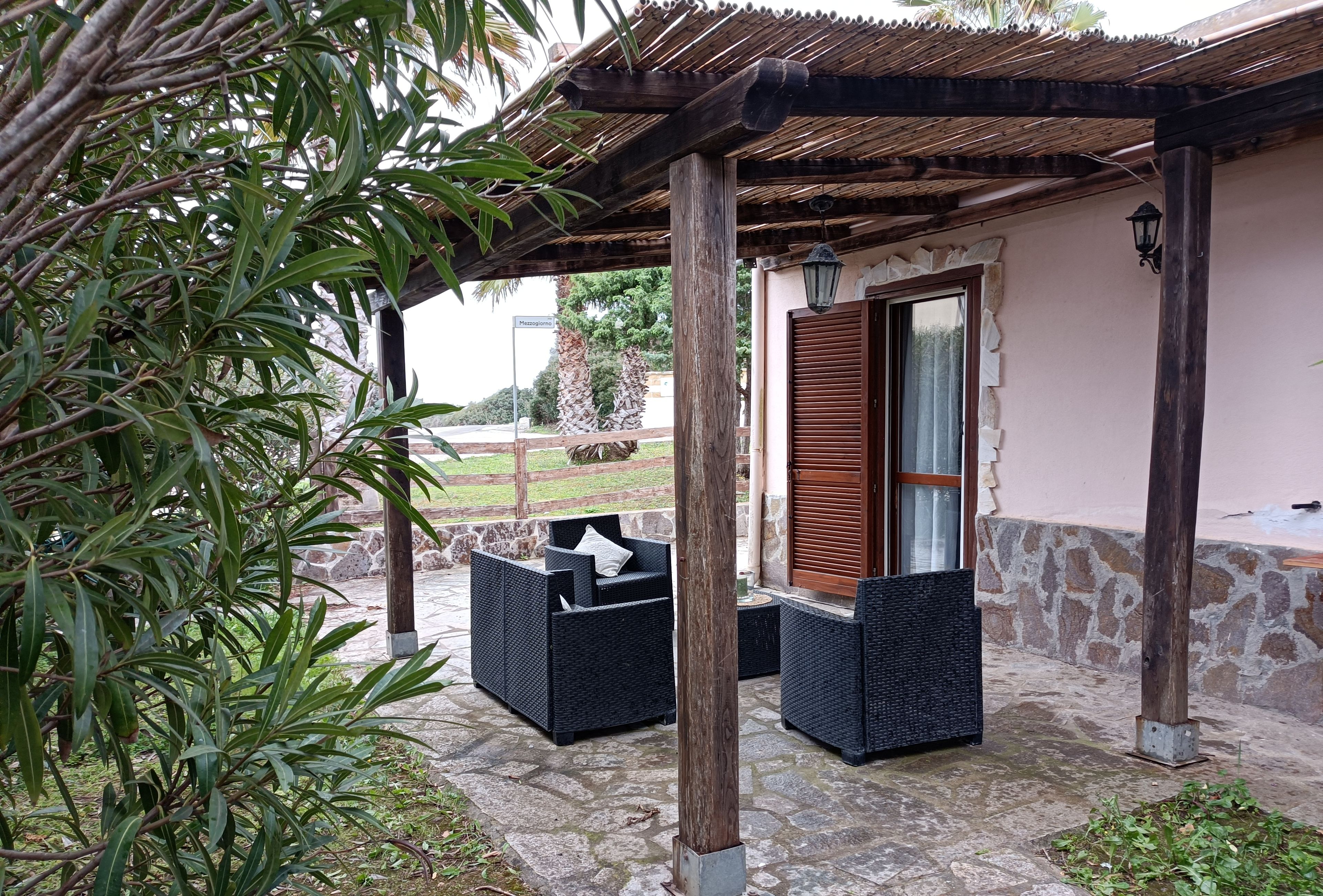 Un área de estar al aire libre con mobiliario de mimbre bajo una pérgola de madera, suelo de piedra y pared de piedra.