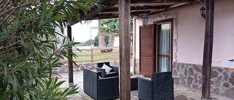 An outdoor seating area with wicker furniture under a wooden pergola, stone flooring, and a stone wall.