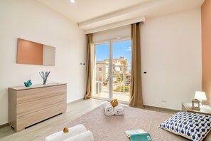 A modern bedroom with a large bed, a wooden dresser, a nightstand, and a view of buildings through a glass door.