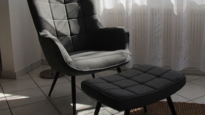 Un salon moderne avec un fauteuil et un pouf gris, une lampe sur pied et une vue à travers des rideaux transparents.