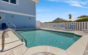 Una piscina azul cristalina con una escalera metálica, rodeada por una cerca blanca y una silla azul.