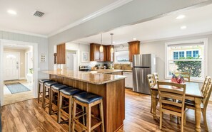 Una cocina amplia con una isla central, taburetes de bar de madera, una mesa de comedor con sillas y un refrigerador de acero inoxidable.