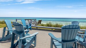 Een terras met blauwe Adirondack-stoelen met uitzicht op een strand en helder turquoise water.