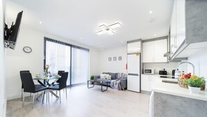 A modern kitchen with white cabinets, a microwave, and a sink. There is a dining area with a glass table and chairs, and a sofa in the living room.