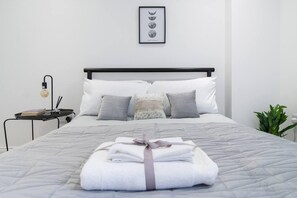A neatly made bed with a grey quilt, multiple pillows, a framed lunar phases picture on the wall, a small side table with a lamp, and a plant by the window.