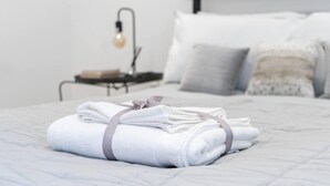 A neatly made bed with folded white towels and a grey ribbon, a bedside table with a lamp, and a bed with grey and white pillows.