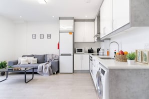 A modern kitchen with white cabinets, a microwave, and a sink. There is a gray sofa with cushions and a small table with plants.
