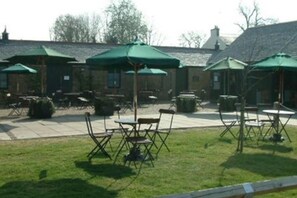 An outdoor seating area with tables and chairs under green umbrellas, a grassy lawn, and a building with a brick facade in the background.