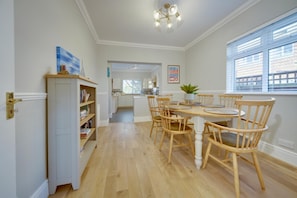 A dining area with a wooden table and chairs, a kitchen in the background, and a view of the outside through a window.