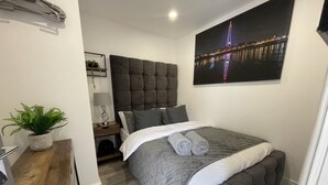 A modern bedroom with a large bed, a headboard, a nightstand, a lamp, a potted plant, and a cityscape painting on the wall. - Osborne Luxury Hot Tub and Jacuzzi Suites (Blackpool)