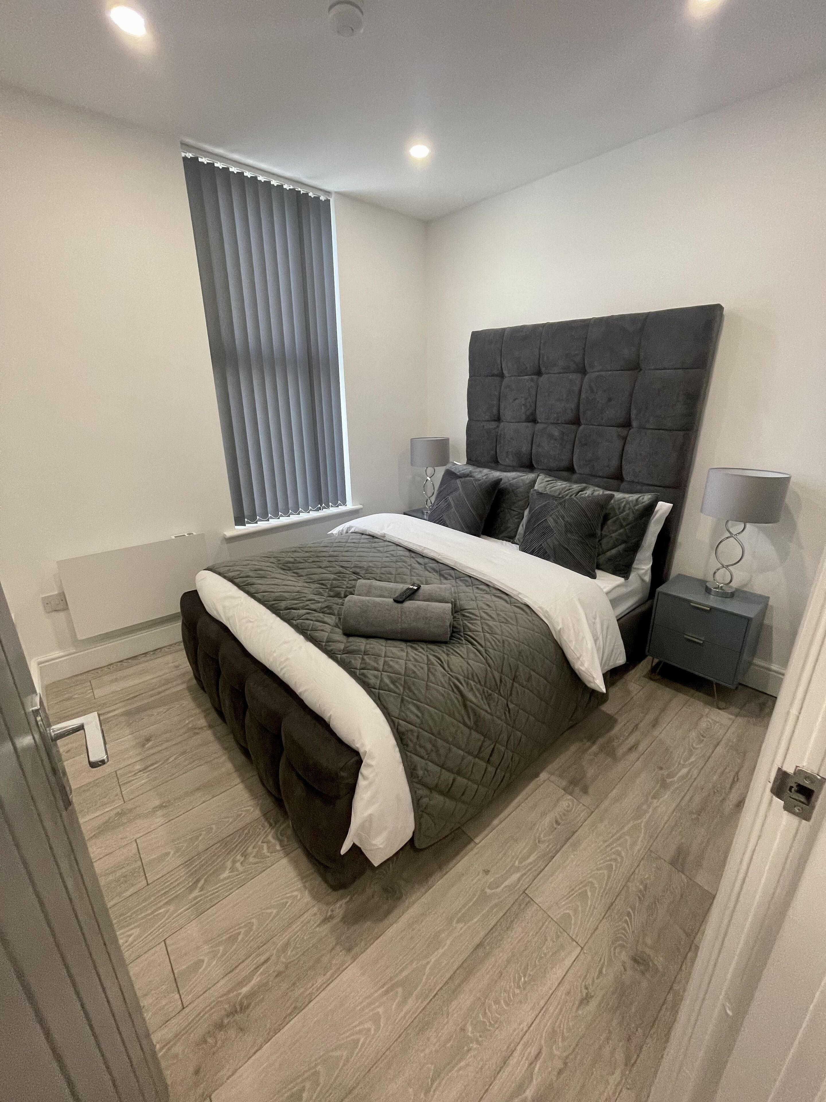 Un dormitorio moderno con una cama grande, cabecero gris, mesitas de noche y una ventana con persianas.