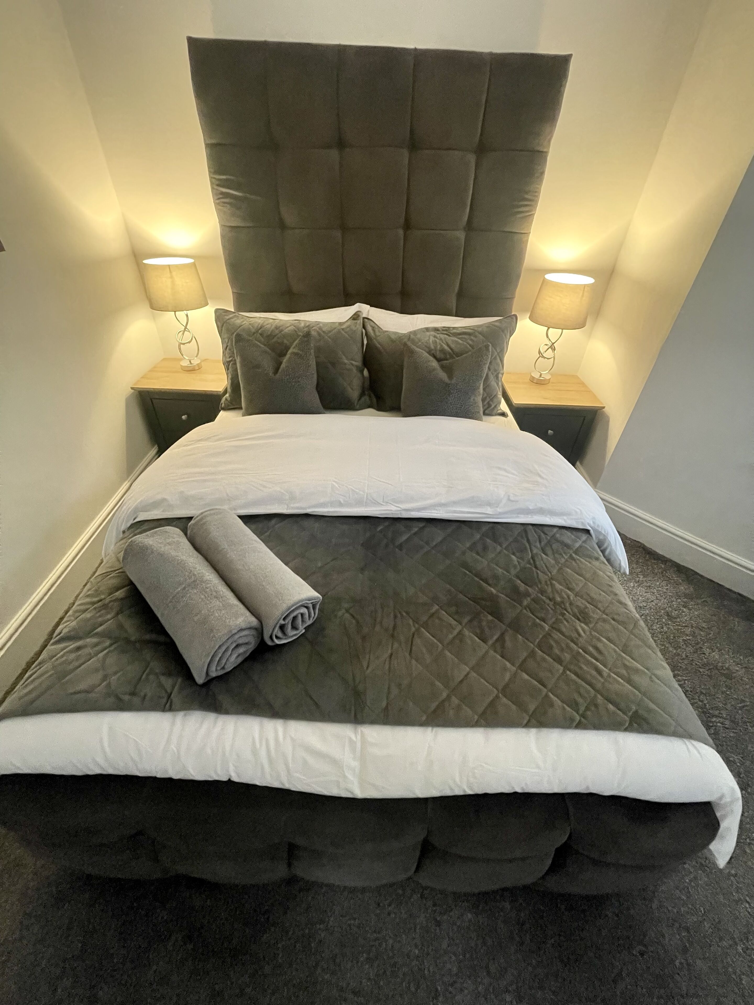 A bed with a tufted headboard, two bedside tables with lamps, and a rolled towel on the bed.
