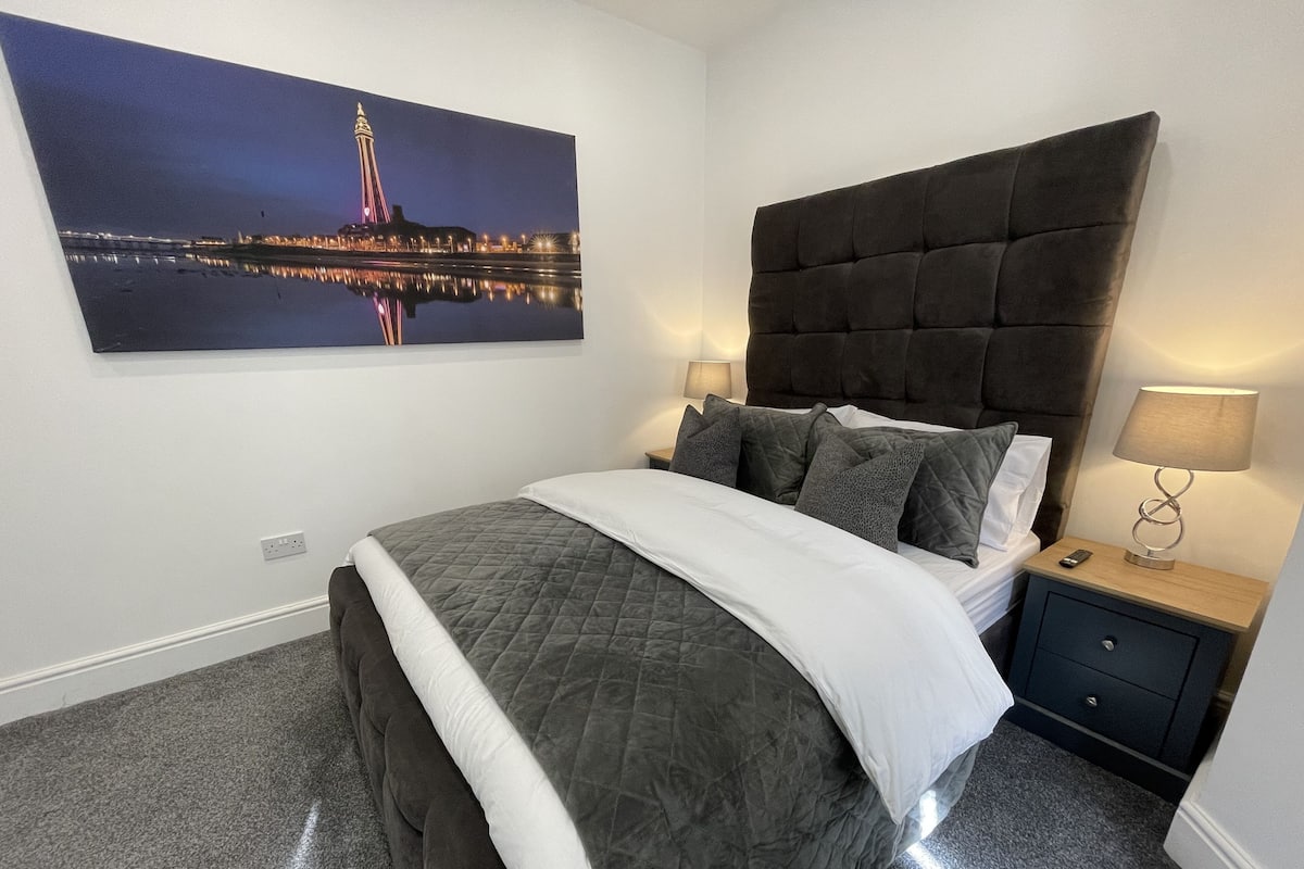 Un dormitorio con una cama grande, una mesita de noche con lámpara, un cajón y un cuadro de un paisaje urbano colgado en la pared.