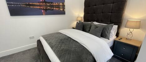 Een slaapkamer met een groot bed, een nachtkastje met een lamp, een lade en een schilderij van een stadsgezicht aan de muur.