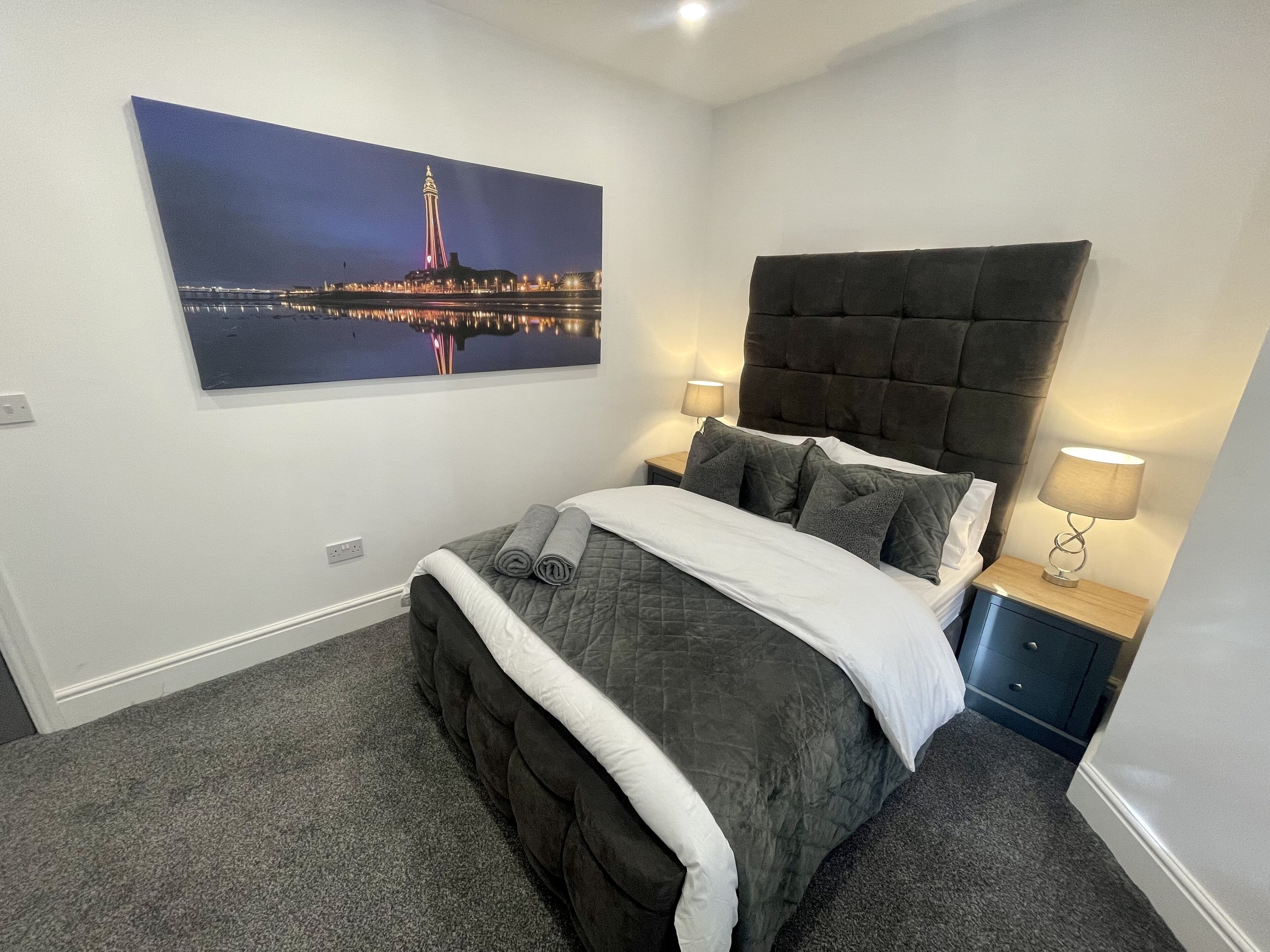 Een net opgemaakt bed met een donker hoofdbord, twee bedlampjes en een stadsgezichtschilderij aan de muur.