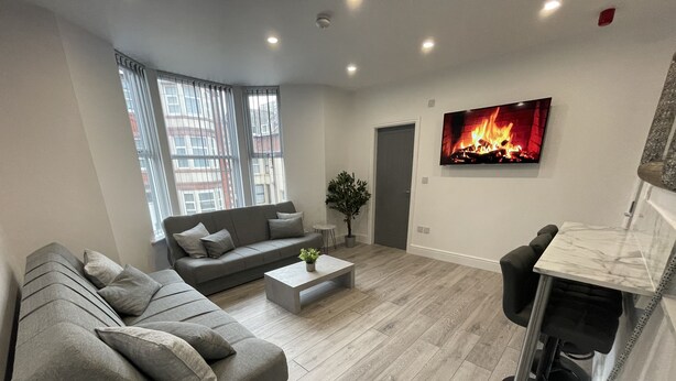 A modern living room with a grey sectional sofa, a coffee table, and a TV displaying a fireplace. - Osborne Luxury Hot Tub and Jacuzzi Suites (Blackpool)
