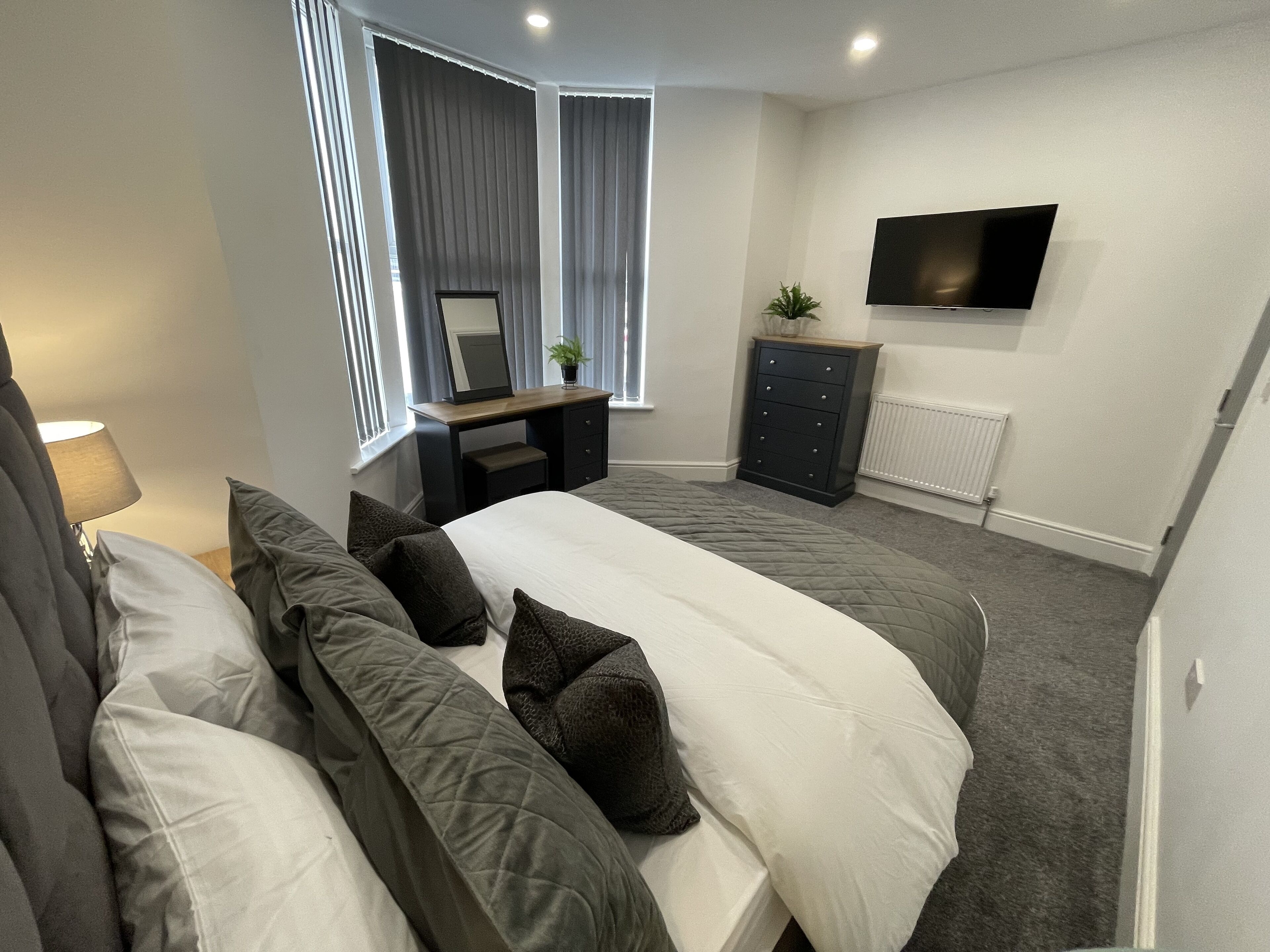Une chambre à coucher moderne avec un grand lit, une commode, un téléviseur fixé au mur et une fenêtre avec des rideaux.