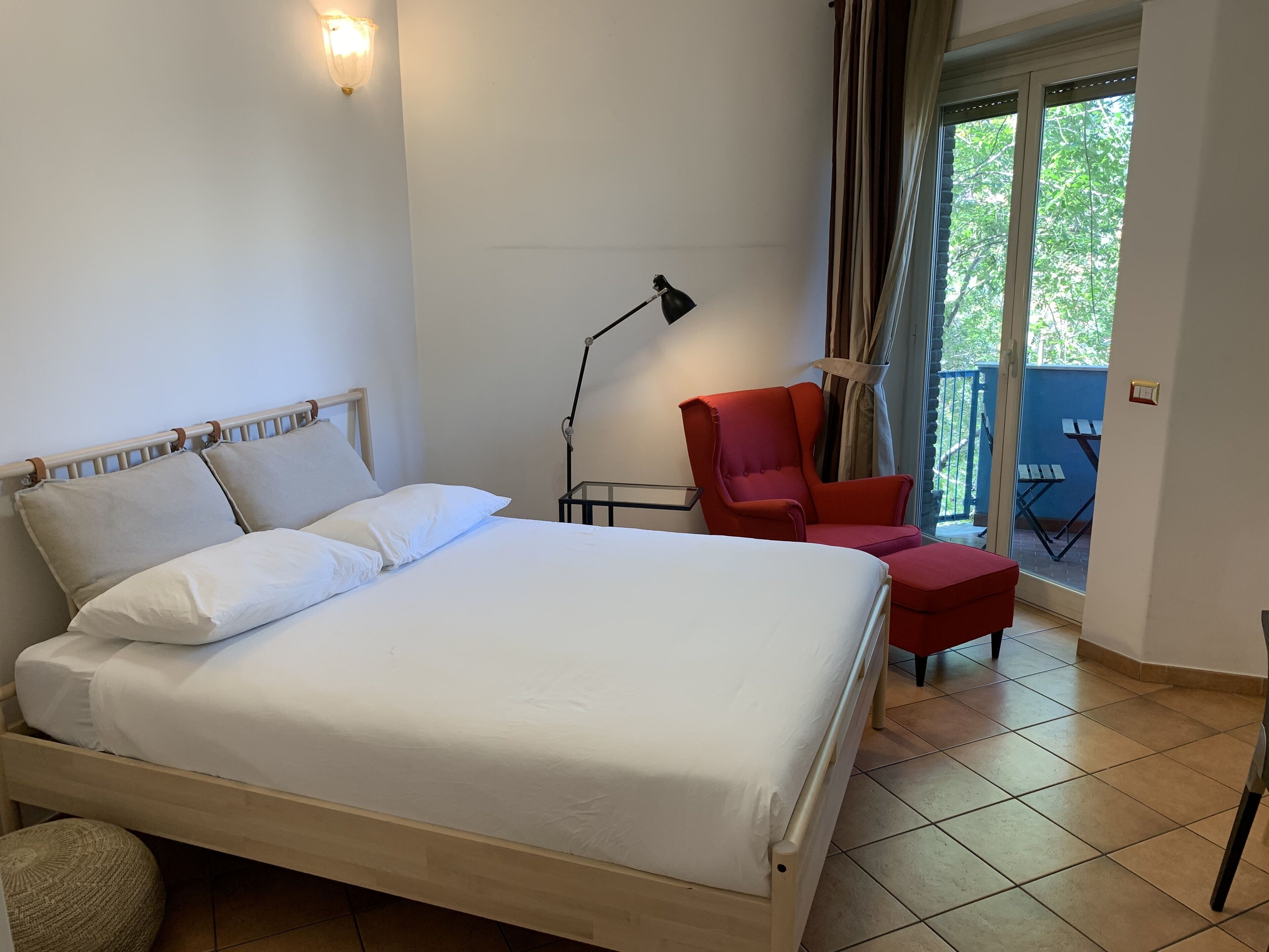 A hotel room with a bed, a red armchair, a small table, a lamp, and a view of the outdoors through a sliding door.