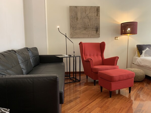 A modern living room with a black leather sofa, a red armchair, a small table, a floor lamp, and a bed with a white comforter. - SmooRoomS Trastevere (Rome)