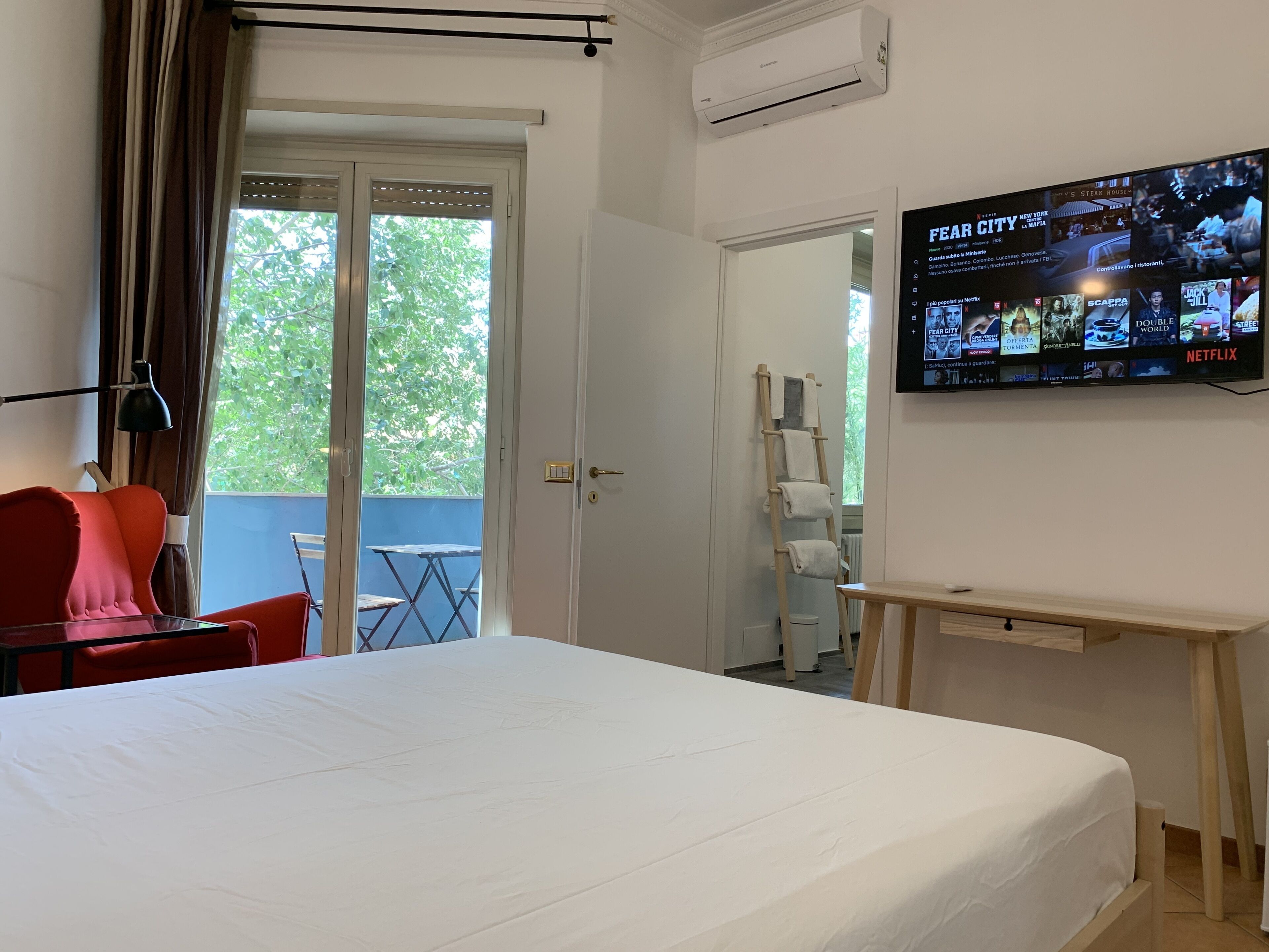 A hotel room with a bed, a desk, a TV displaying a movie, a red chair, a table, and a view of the outdoors through a sliding door.