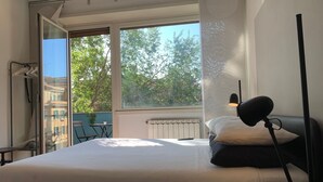 A bedroom with a bed, a nightstand, a lamp, a radiator, and a view of trees through large windows. - SmooRoomS Trastevere (Rome)