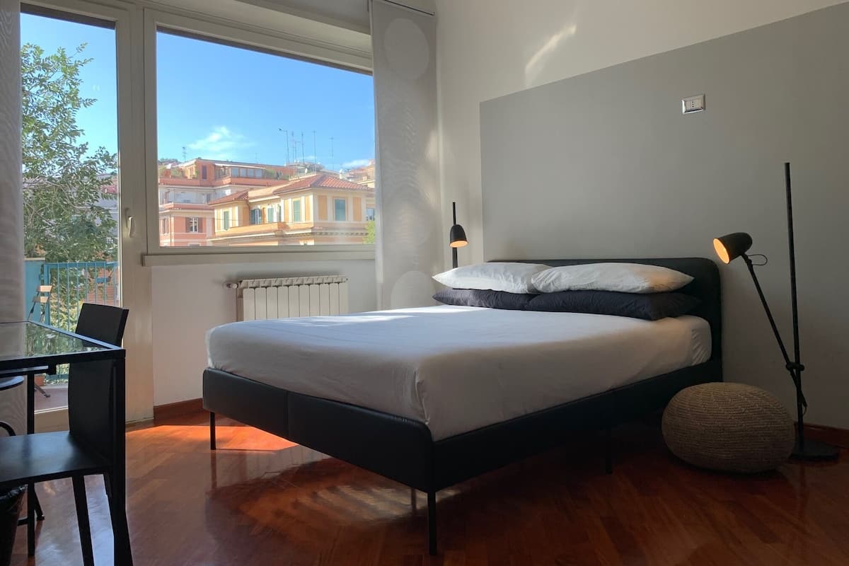 A bedroom with a bed, a desk, and a chair. There is a window with a view of buildings outside.