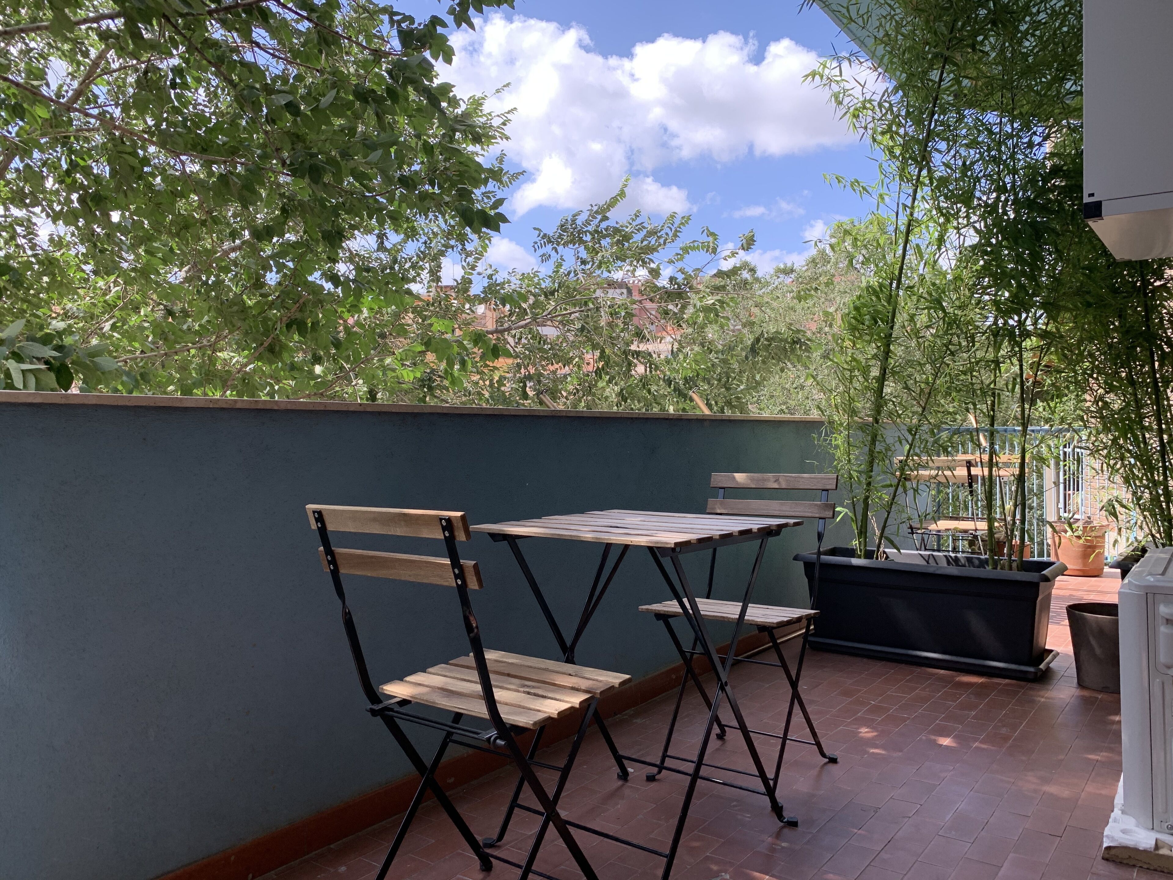 A patio with two wooden tables and chairs, a potted plant, and a view of trees and buildings.