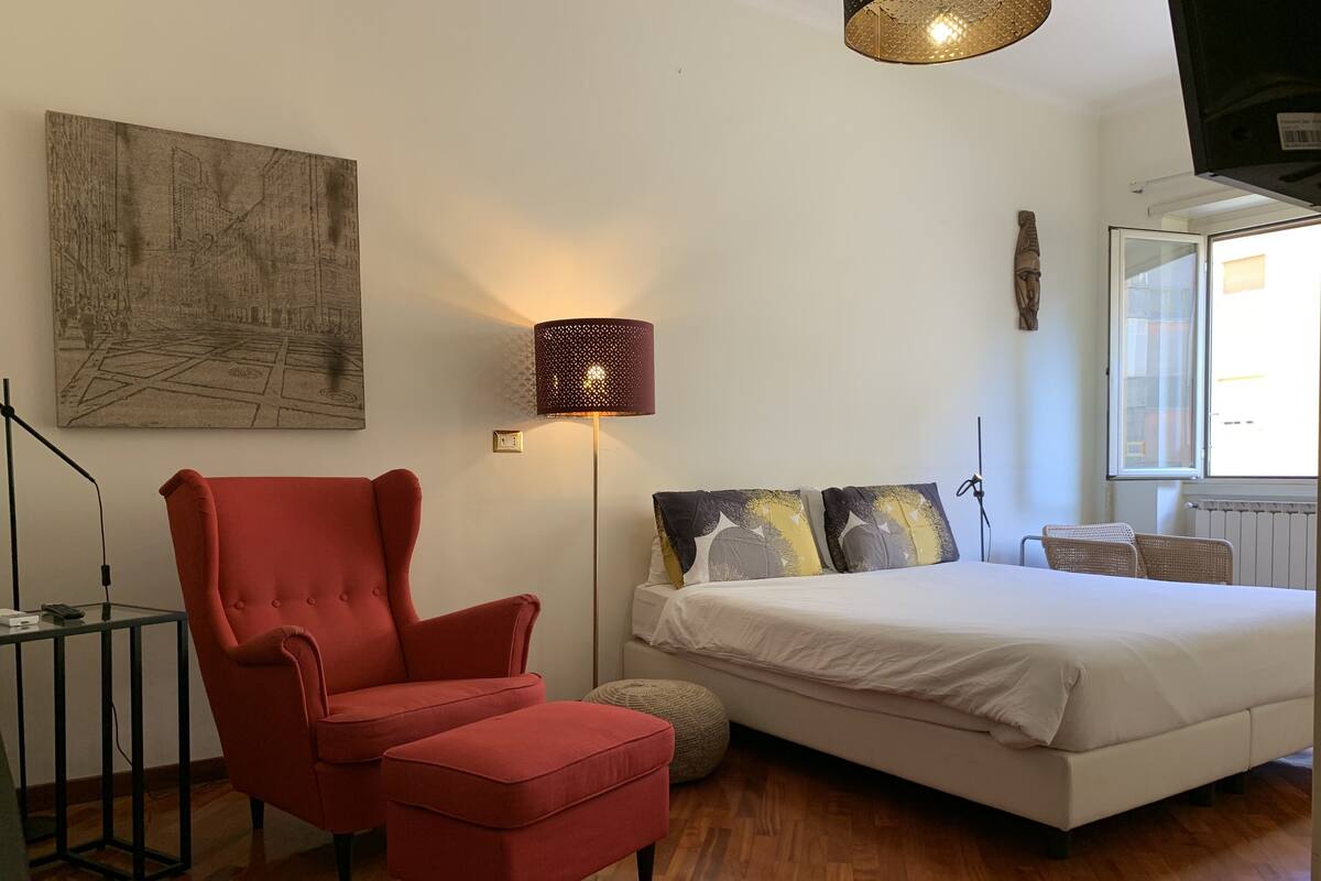 A bedroom with a red armchair, a bed with patterned pillows, a standing lamp, and a large abstract painting on the wall.