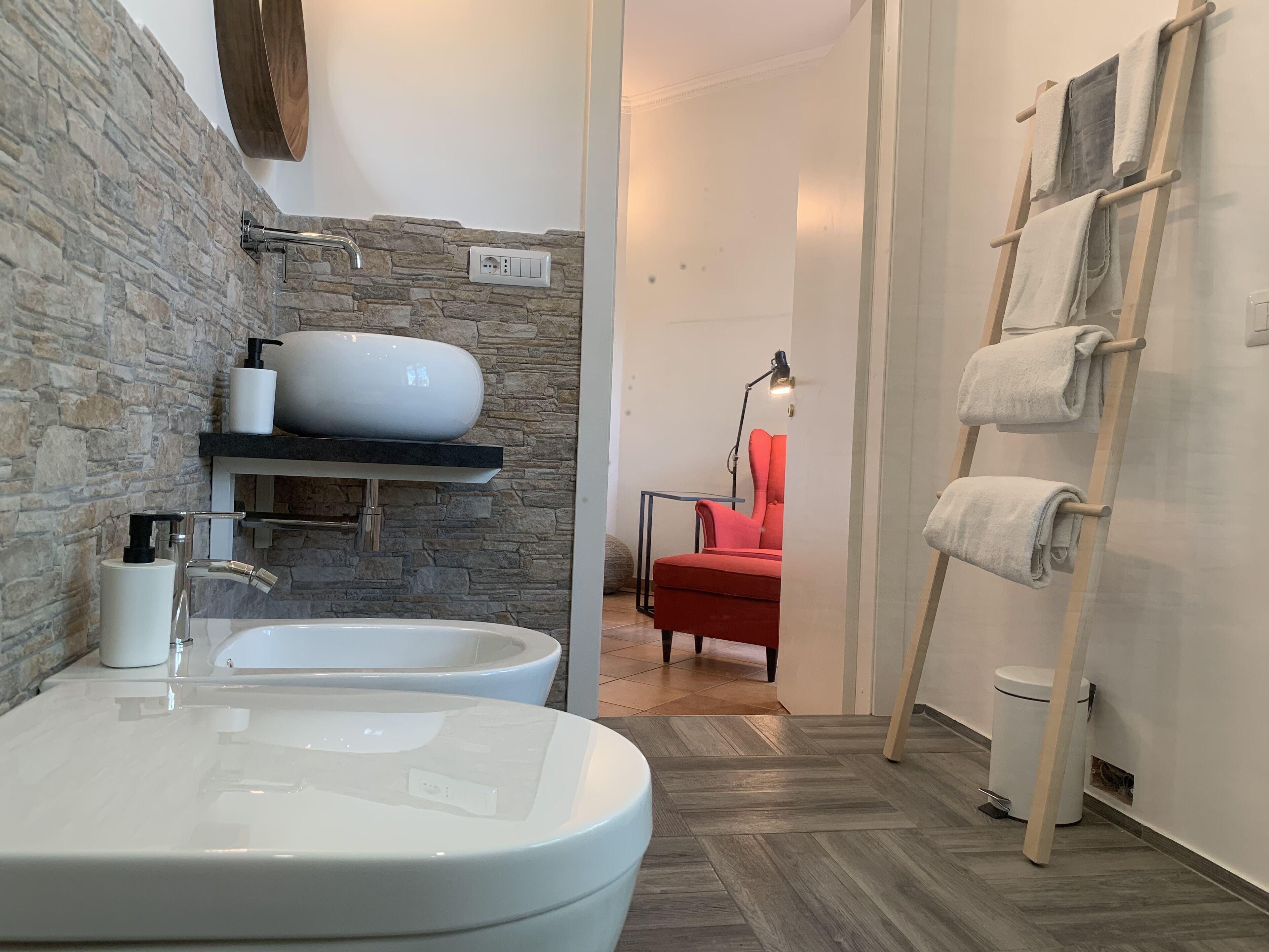 A bathroom with two sinks, a stone accent wall, a red chair, and a towel rack.