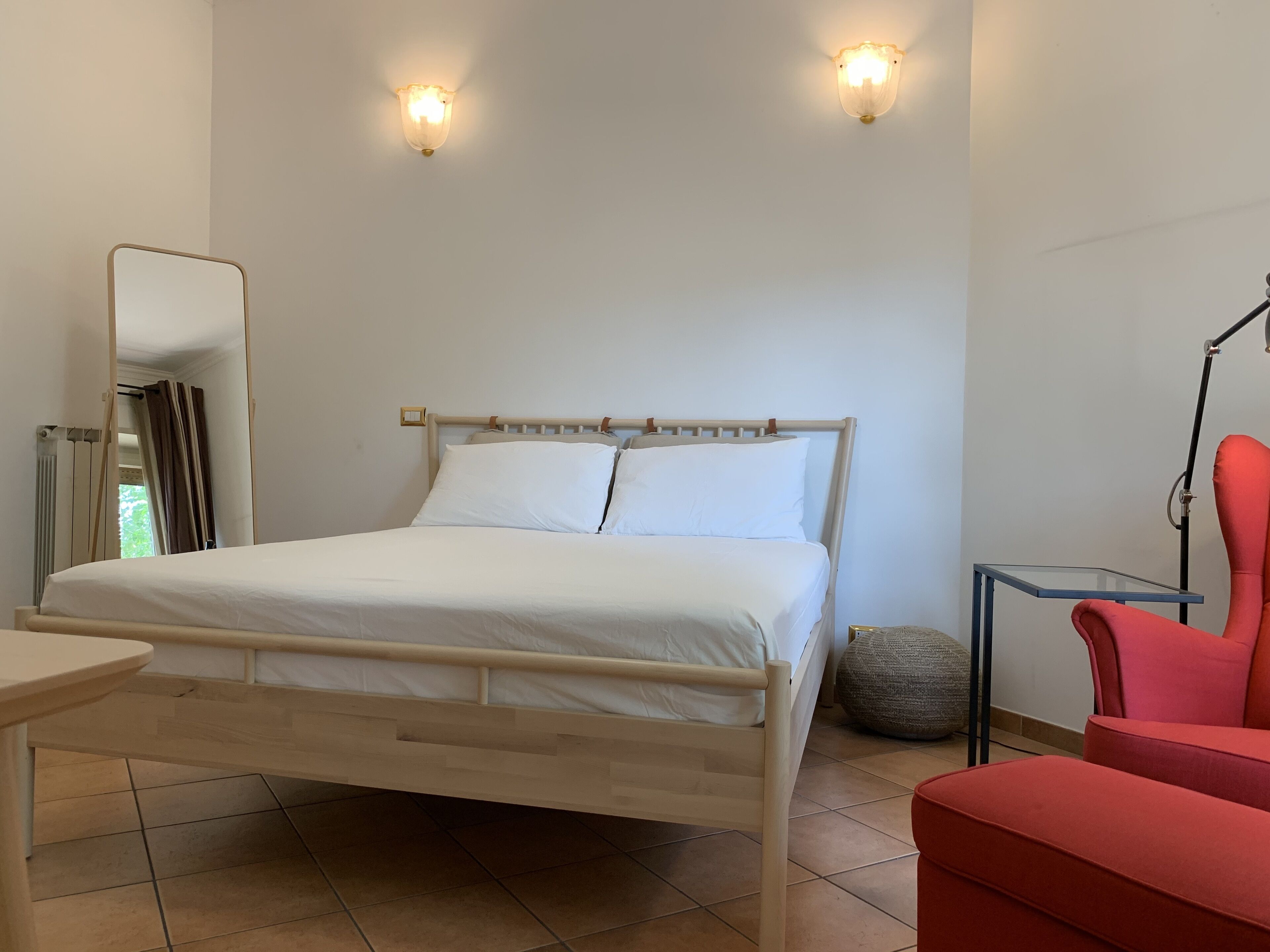 Ein Schlafzimmer mit einem hölzernen Bett, weißer Bettwäsche, einem roten Sessel, einem kleinen Tisch und zwei Wandleuchten.