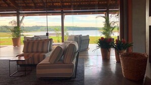 A spacious indoor area with a large window overlooking a lake, furnished with a sofa and chairs, and surrounded by potted plants. - Buganville Villas (Itai)