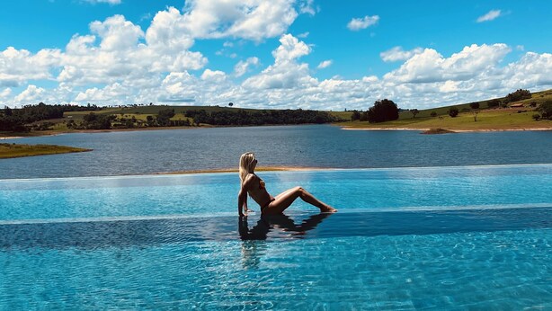 A person sitting on the edge of a clear blue pool with a lake and hills in the background. - Buganville Villas (Itai)