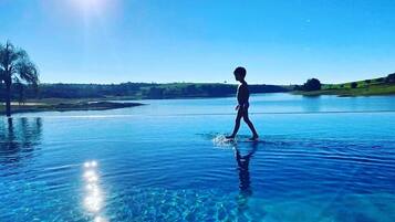 Uma pessoa caminhando em uma piscina azul cristalina, com vista para uma paisagem distante e uma palmeira.