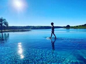 A person walking in a clear blue pool with a view of a distant landscape and a palm tree. - Buganville Villas (Itai)