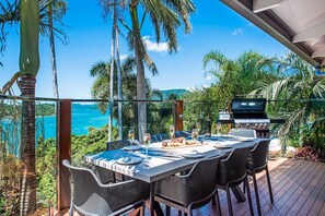 Un espacio para comer al aire libre con vistas al océano, una parrilla y abundante vegetación.