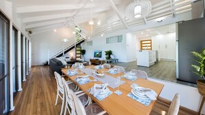 Un comedor moderno con una larga mesa de madera dispuesta para una comida, una escalera que sube a un nivel superior y una cocina visible al fondo.
