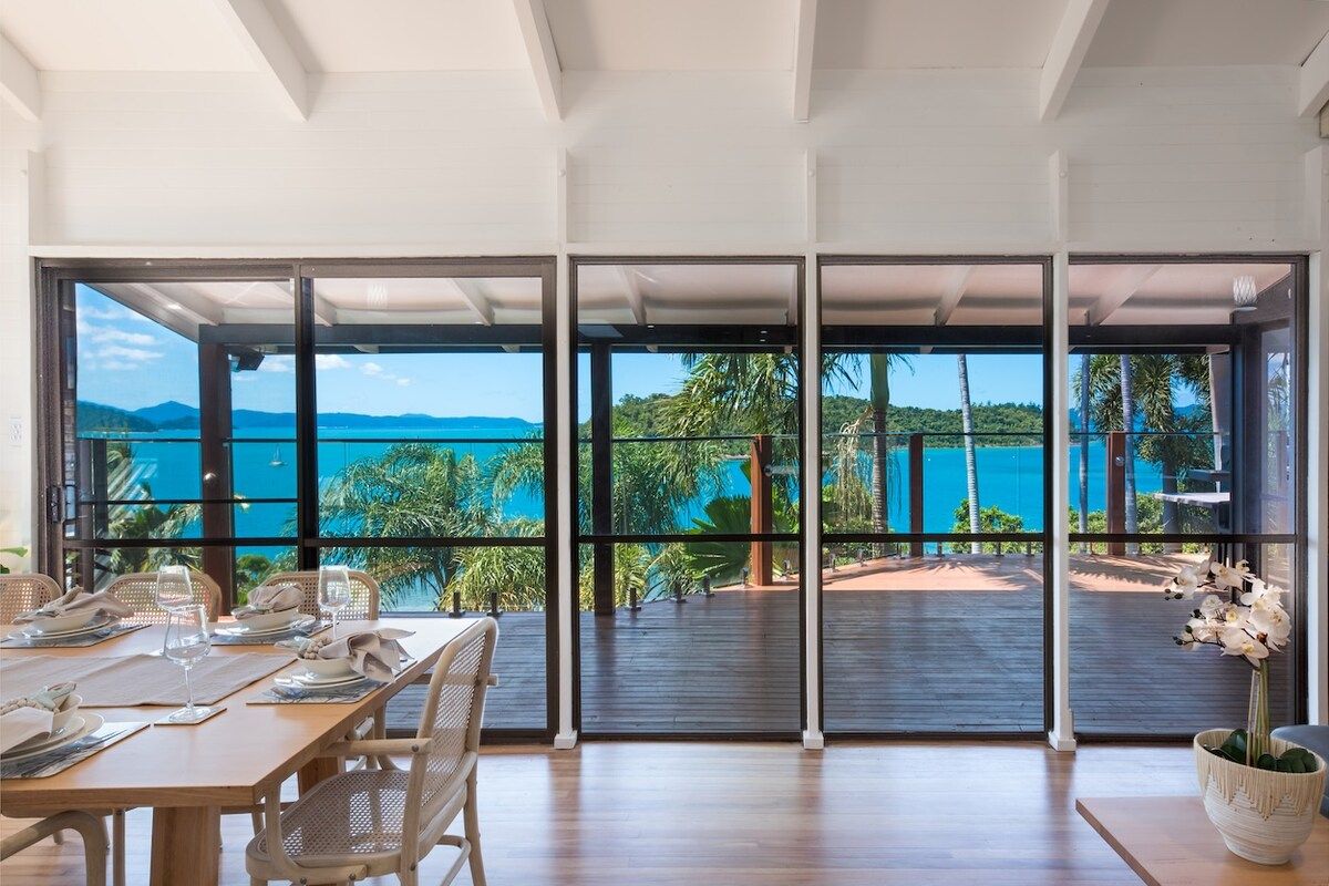 A dining area with a table set for two, large windows offering a view of the sea and greenery.