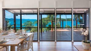 A dining area with a table set for two, large windows offering a view of the sea and greenery. - Coral Sea Pearl (Shute Harbour)